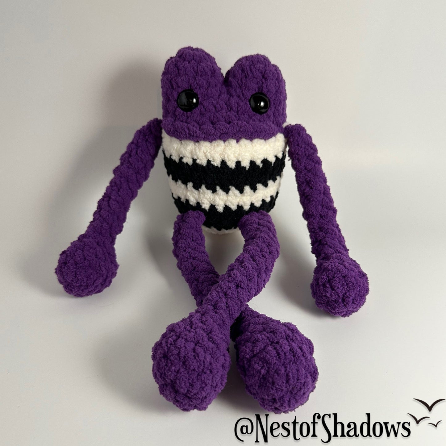 Purple crochet frog on a white background. Nest of Shadows watermark.