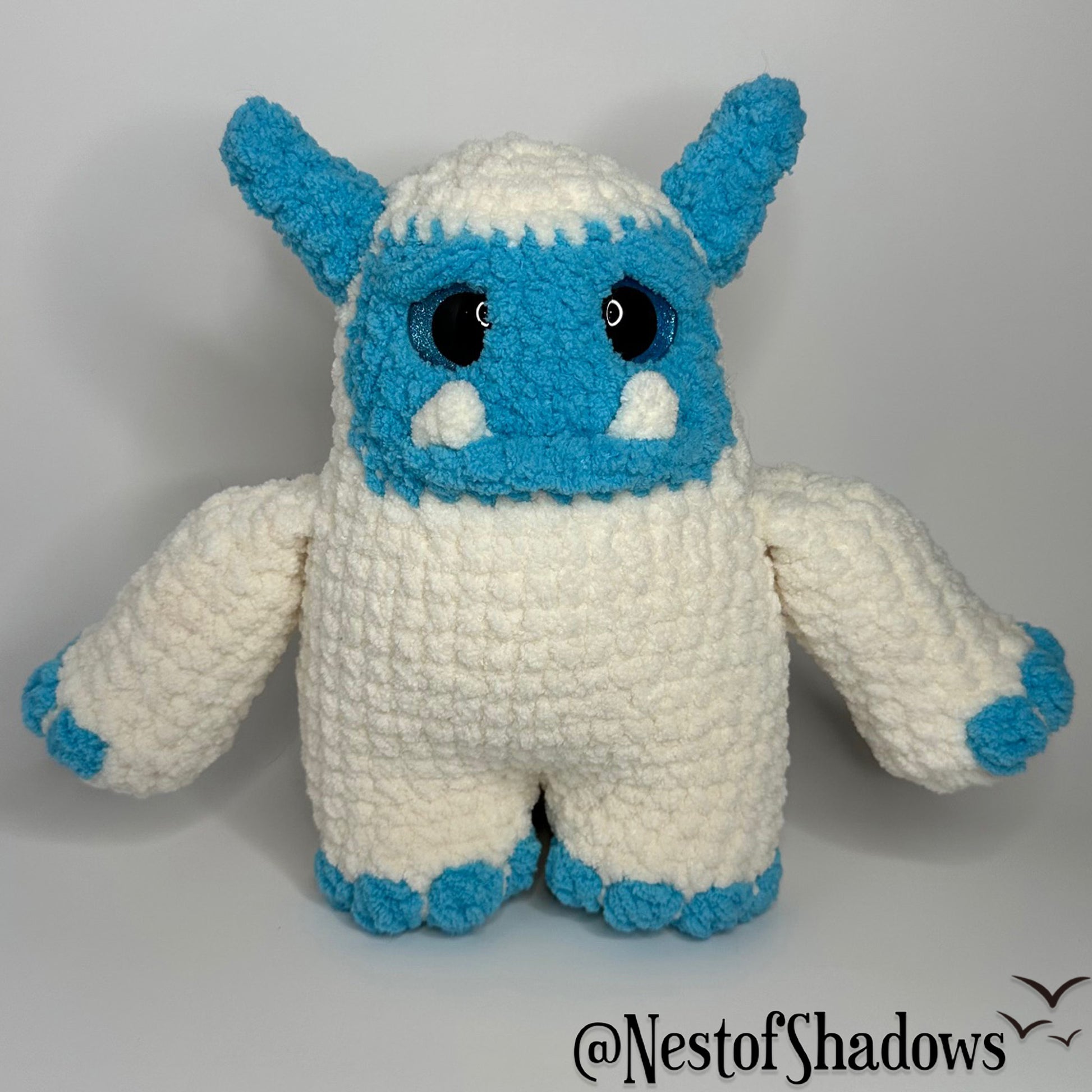Blue and white abominable snowman crochet plushie with a white background and Nest of Shadows watermark