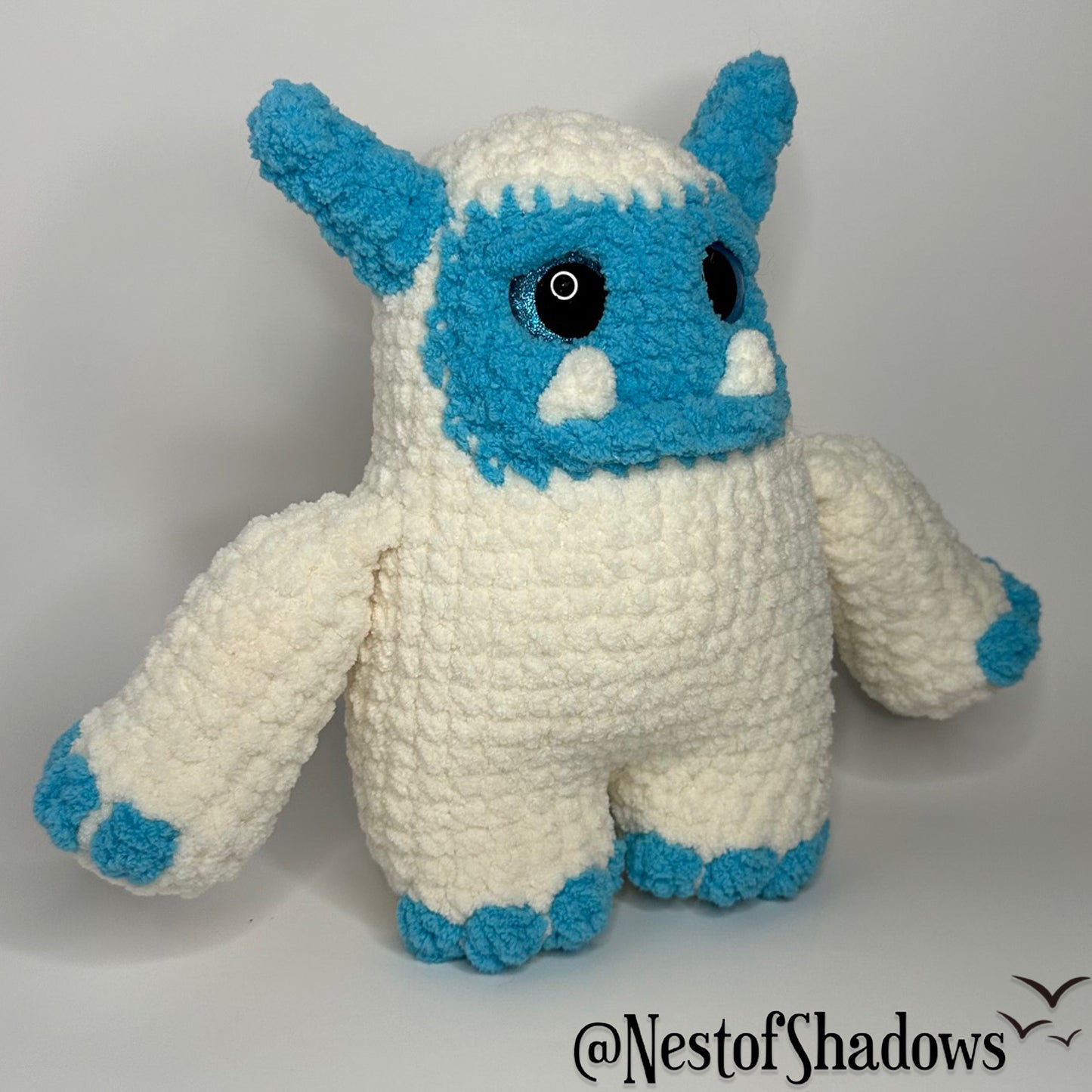 Blue and white abominable snowman crochet plushie with a white background and Nest of Shadows watermark