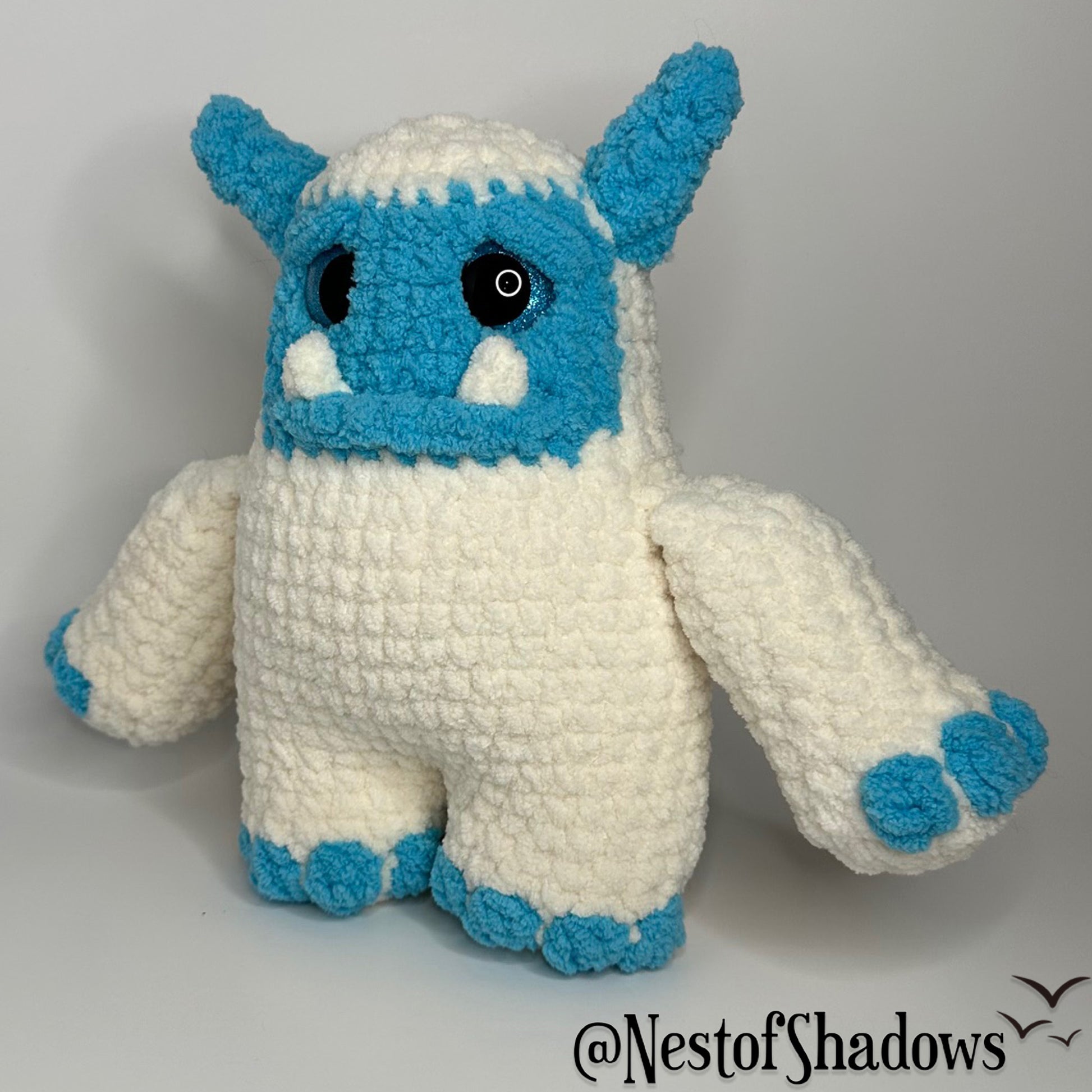 Blue and white abominable snowman crochet plushie with a white background and Nest of Shadows watermark