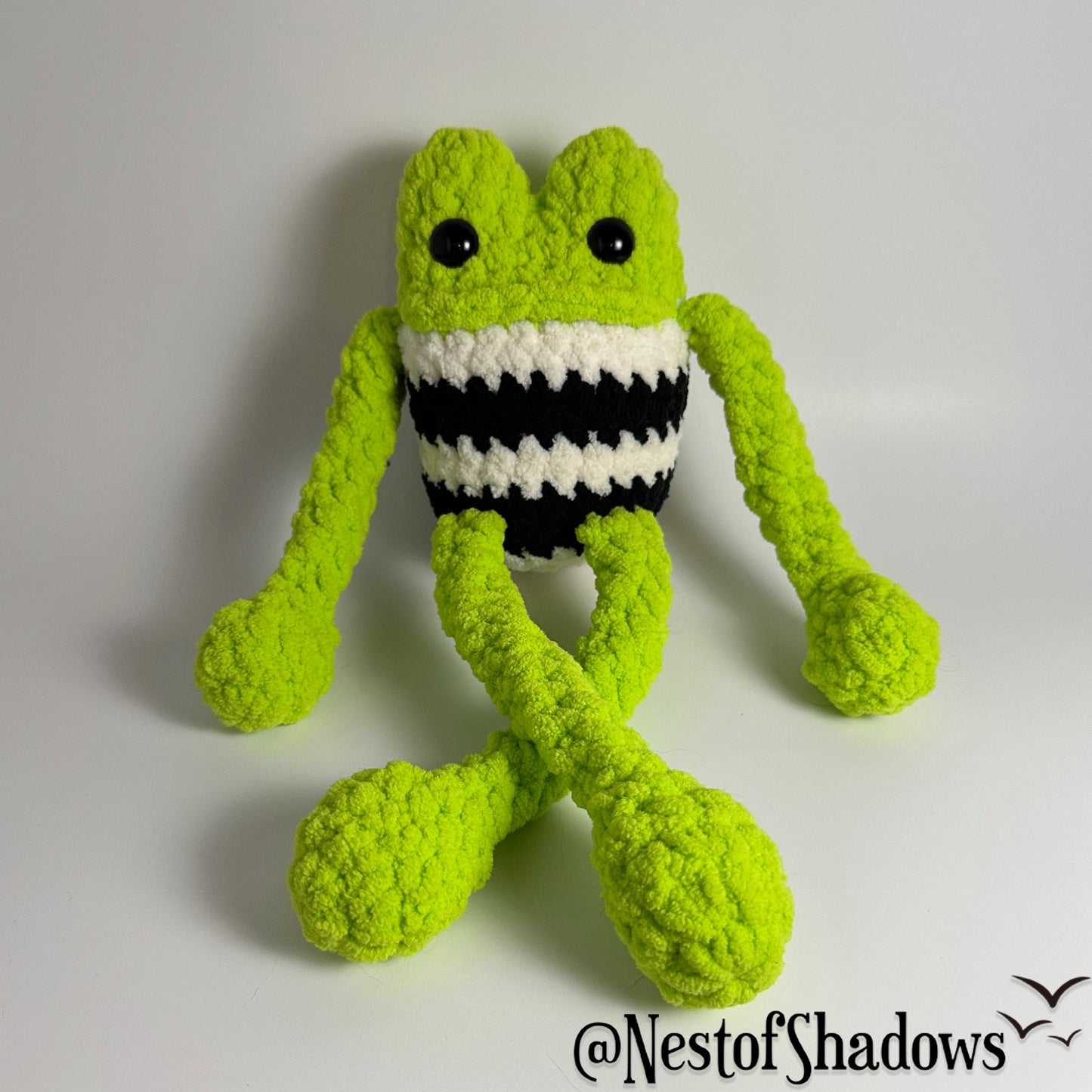 Green crochet frog on a white background. Nest of Shadows watermark.