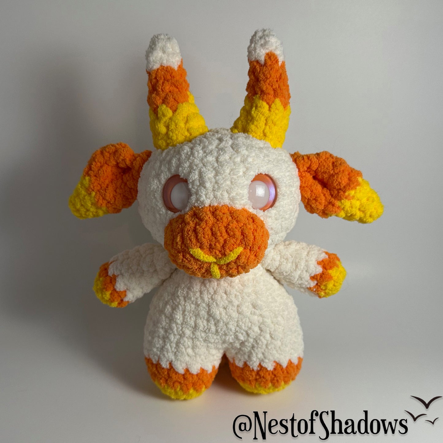 Crochet goat plushie in orange, yellow and white, with a white background and a Nest of Shadows watermark.