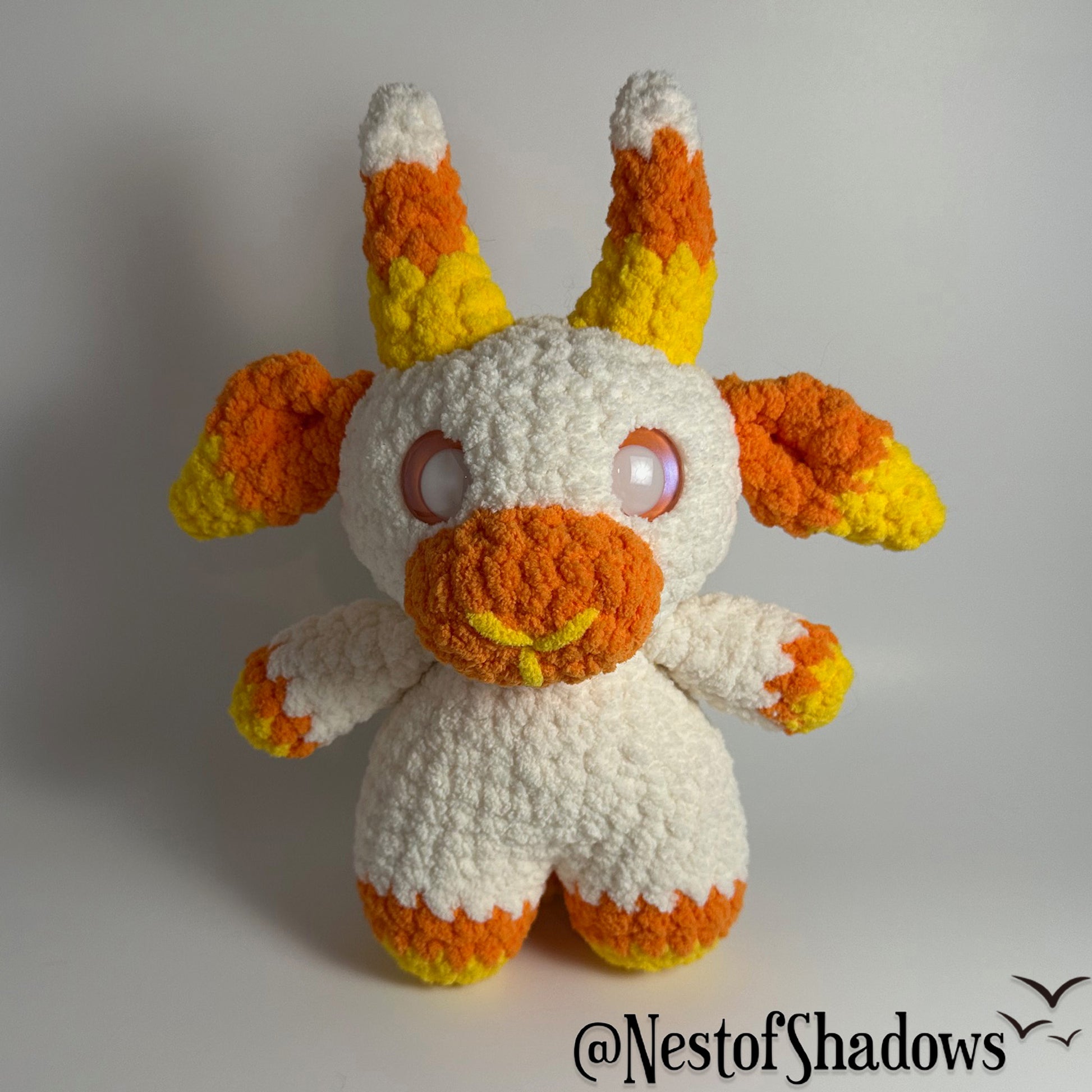 Crochet goat plushie in orange, yellow and white, with a white background and a Nest of Shadows watermark.