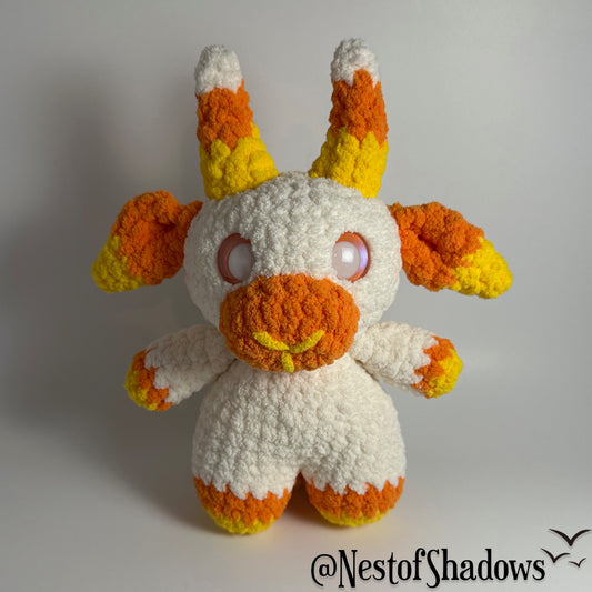 Crochet goat plushie in orange, yellow and white, with a white background and a Nest of Shadows watermark.