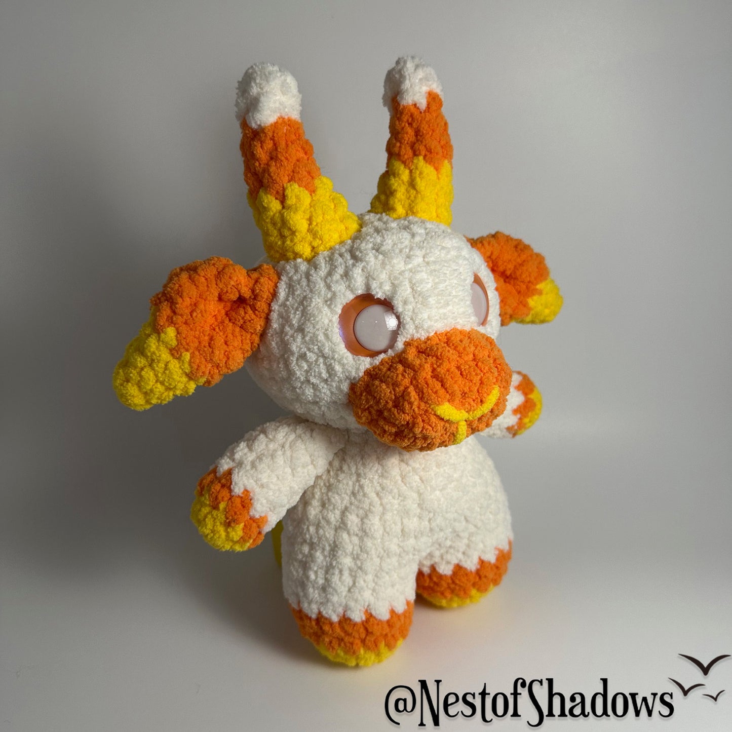 Crochet goat plushie in orange, yellow and white, with a white background and a Nest of Shadows watermark.