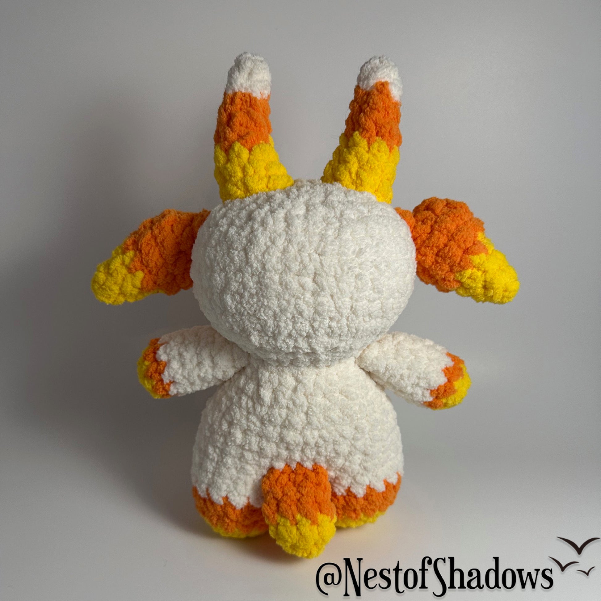 Crochet goat plushie in orange, yellow and white, with a white background and a Nest of Shadows watermark.