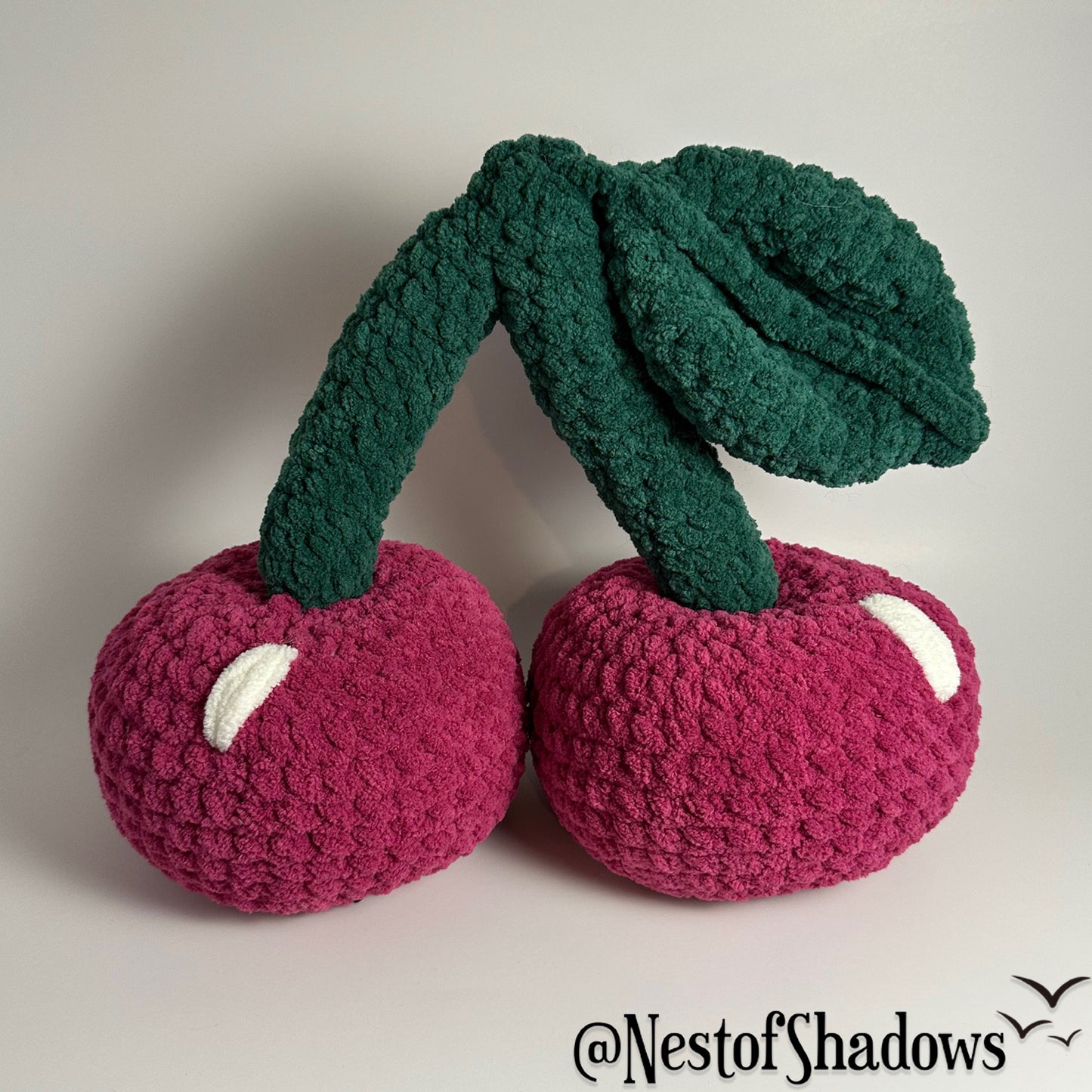 Two crocheted pink cherries on a white background. Nest of Shadows watermark.