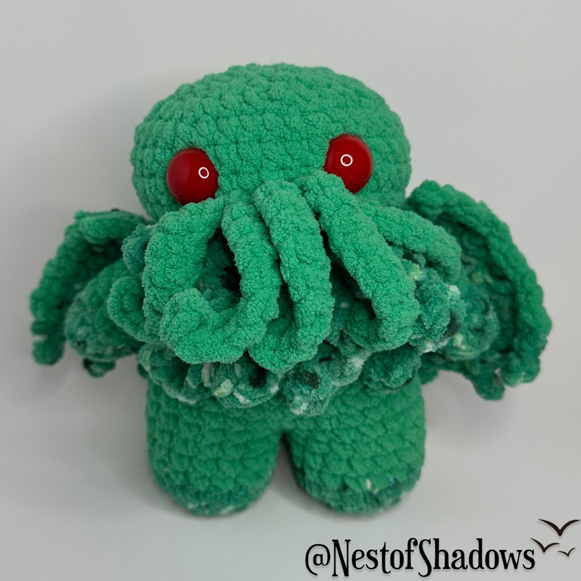 Green crochet cthulhu plushie on a white background. Nest of Shadows watermark.