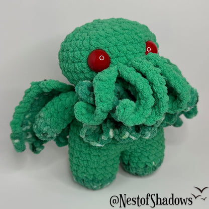 Green crochet cthulhu plushie on a white background. Nest of Shadows watermark.