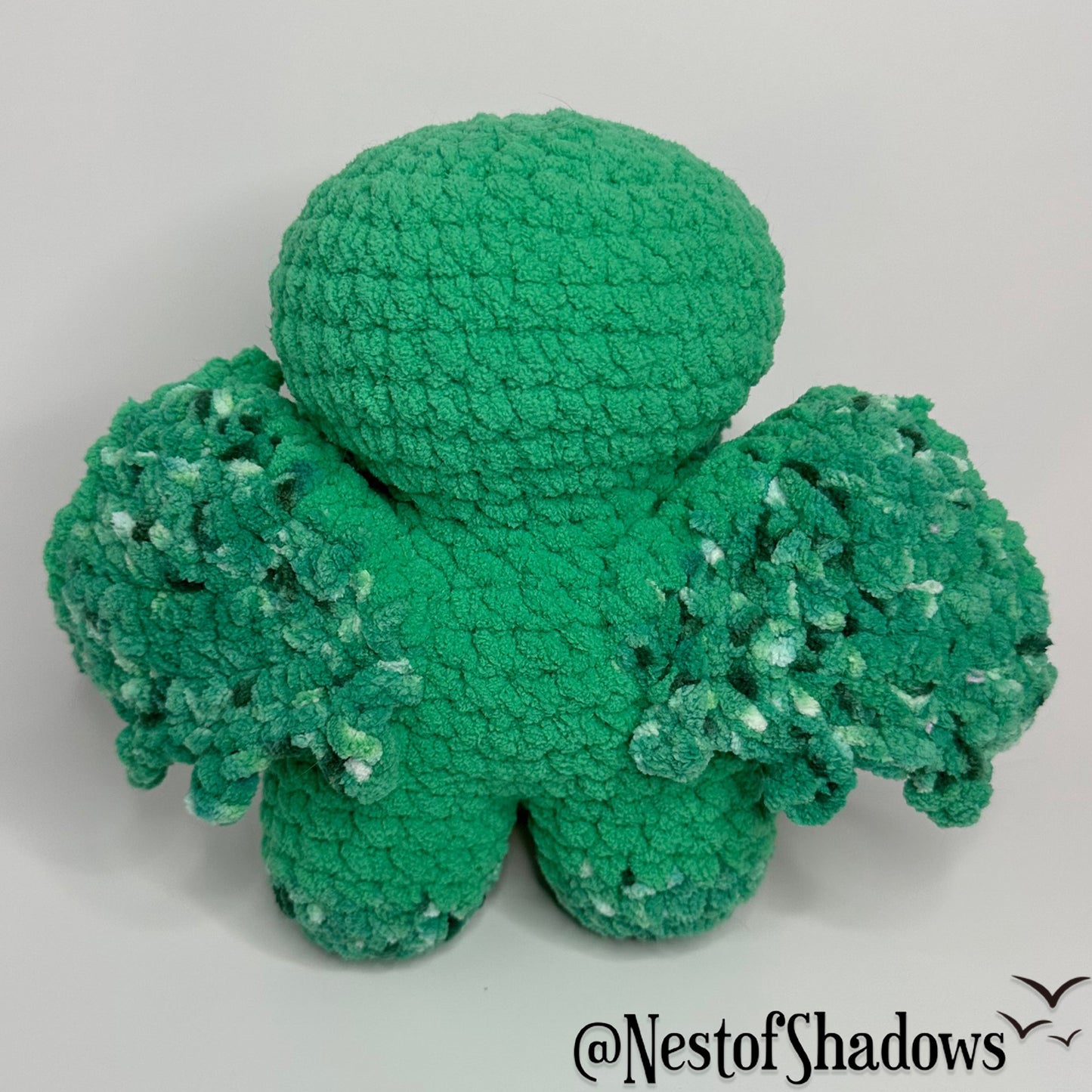 Green crochet cthulhu plushie on a white background. Nest of Shadows watermark.