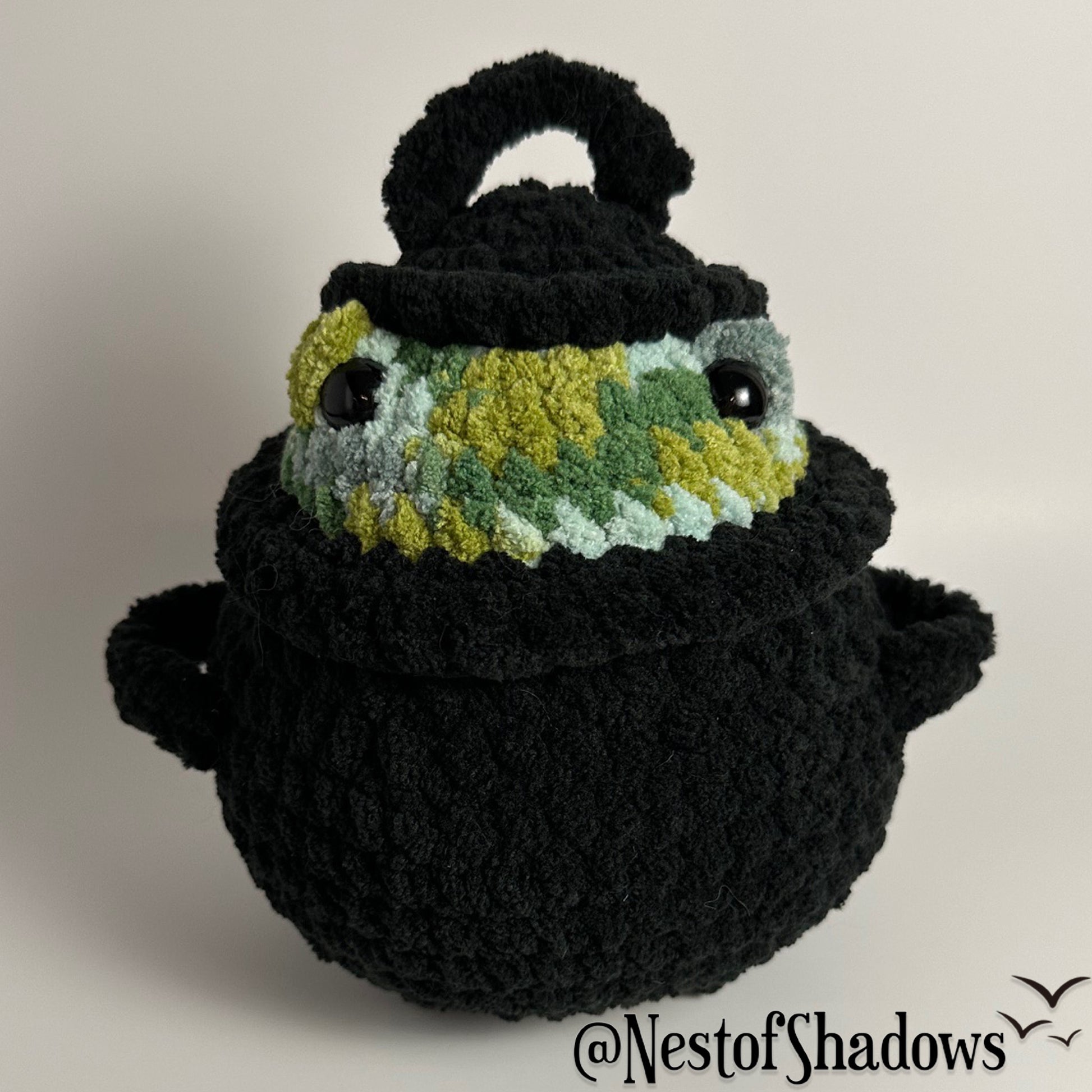 Crochet plushie of frog in a black cauldron on a white background. Nest of Shadows watermark.