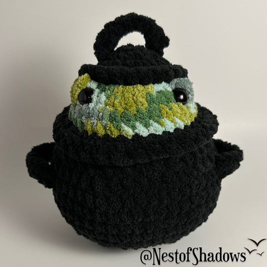 Crochet plushie of frog in a black cauldron on a white background. Nest of Shadows watermark.