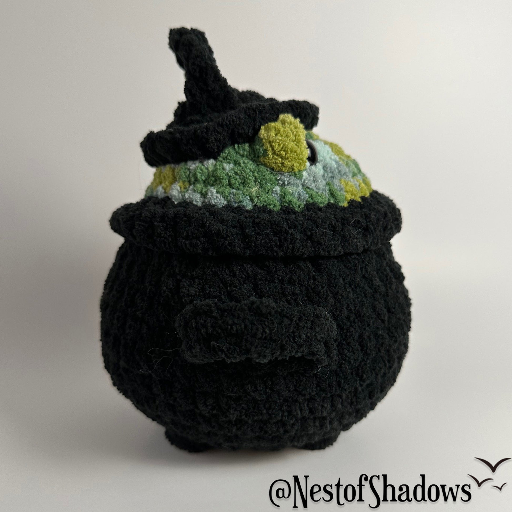Crochet plushie of frog in a black cauldron on a white background. Nest of Shadows watermark.