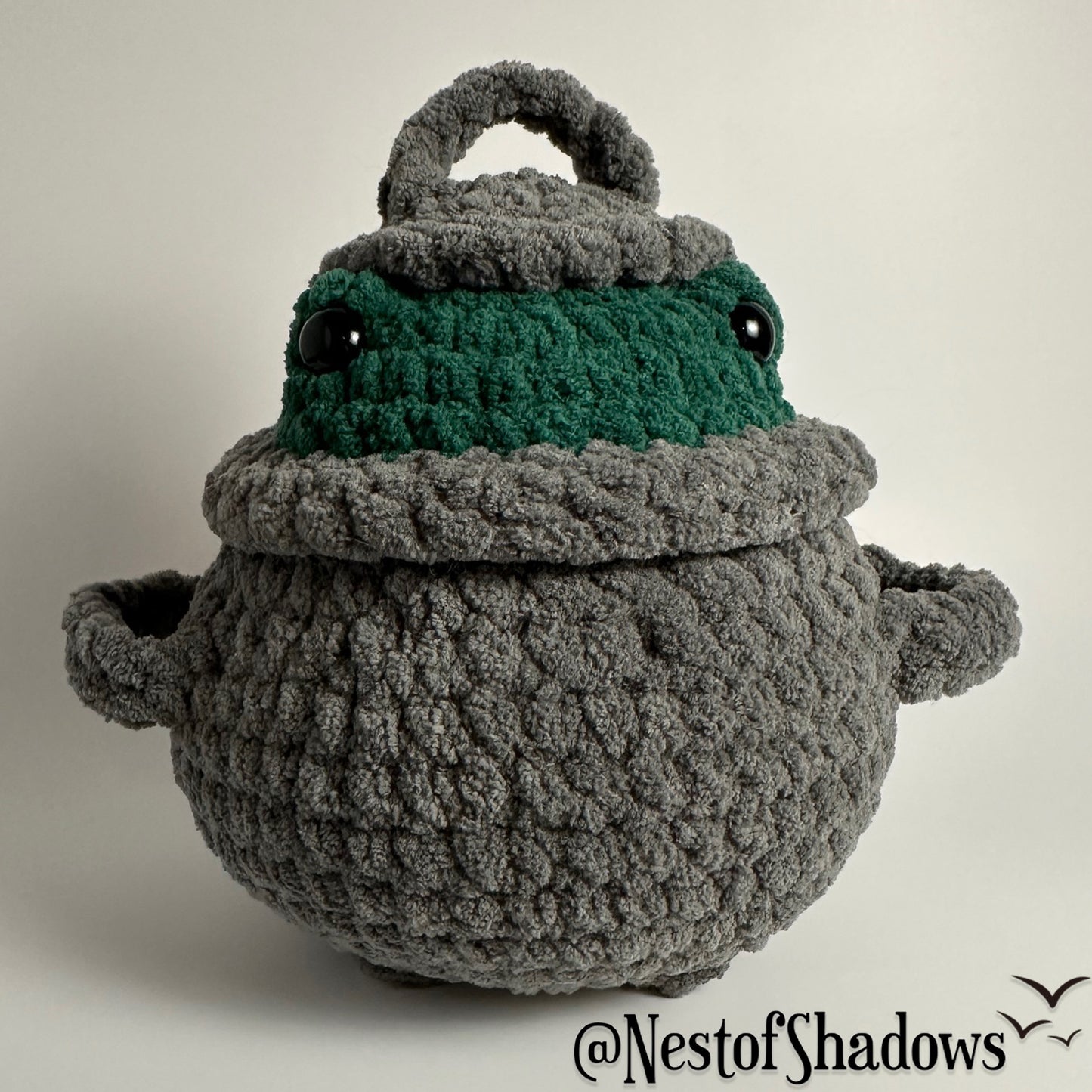 Crochet plushie of frog in a gray cauldron on a white background. Nest of Shadows watermark.