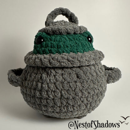 Crochet plushie of frog in a gray cauldron on a white background. Nest of Shadows watermark.