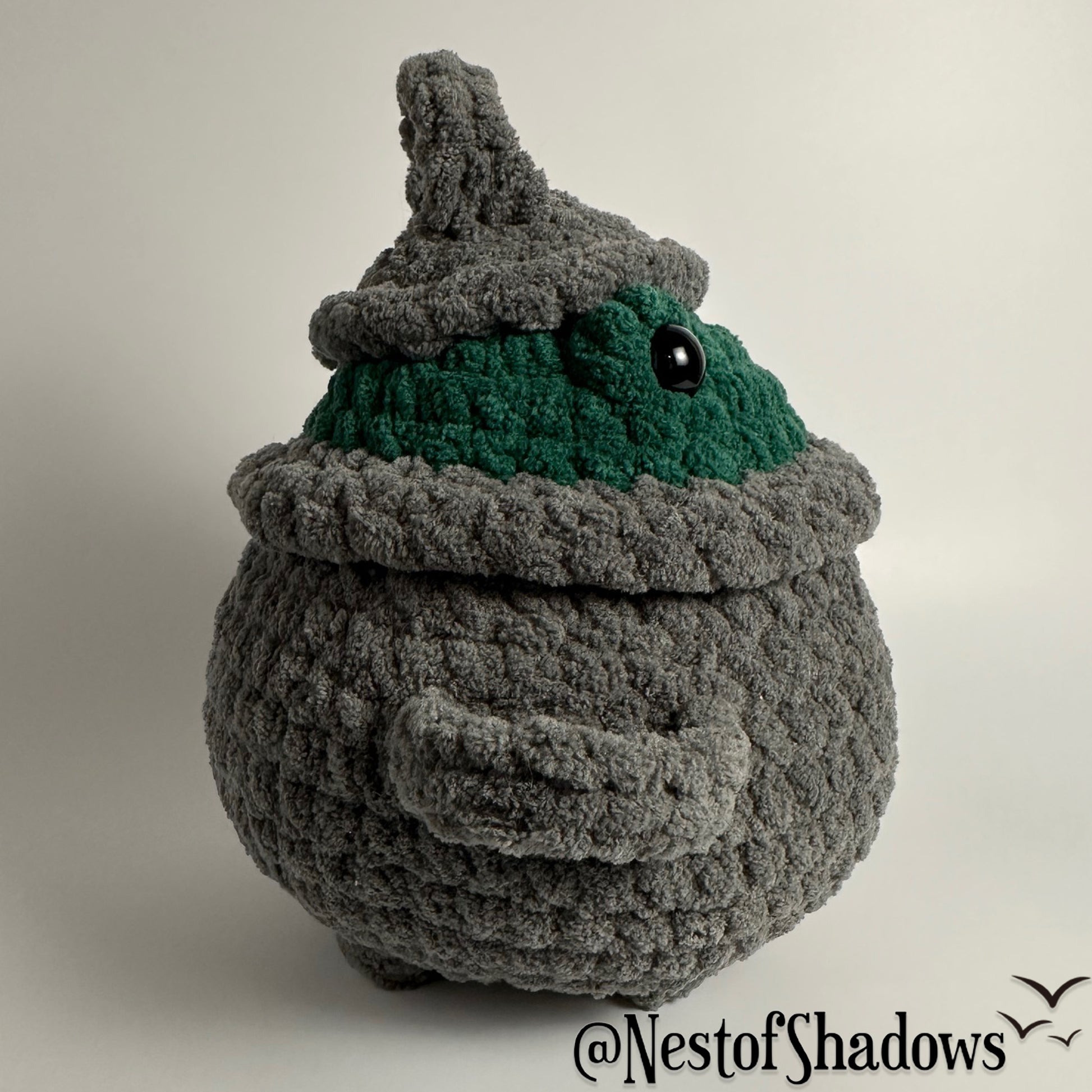 Crochet plushie of frog in a gray cauldron on a white background. Nest of Shadows watermark.
