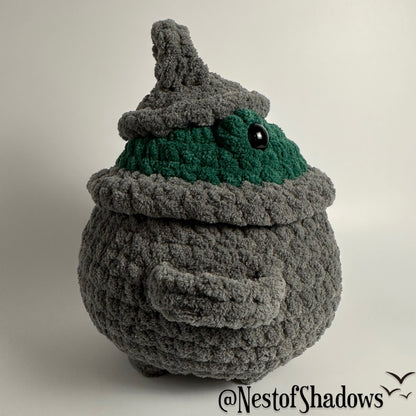 Crochet plushie of frog in a gray cauldron on a white background. Nest of Shadows watermark.