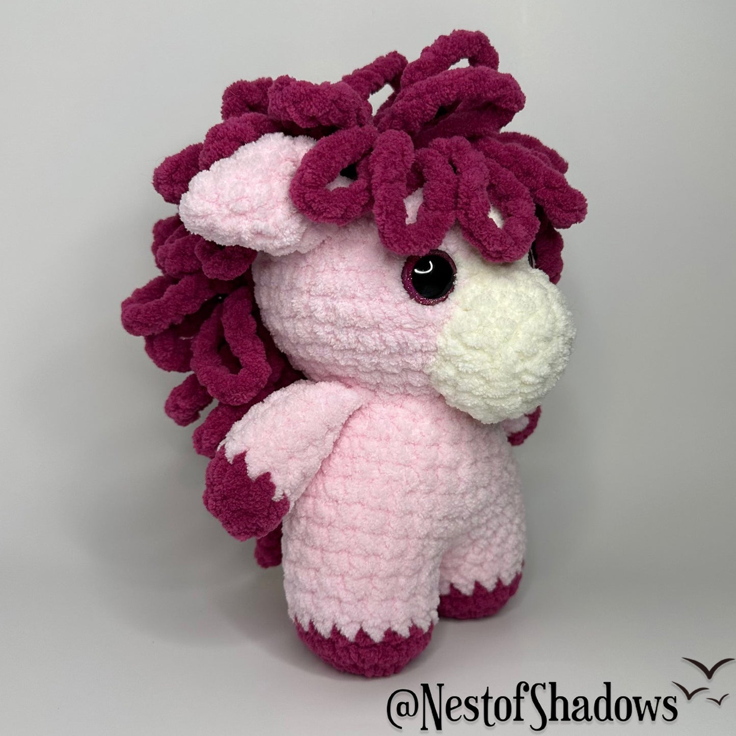 Pink pony horse crochet plushie with a white background a Nest of Shadows watermark.