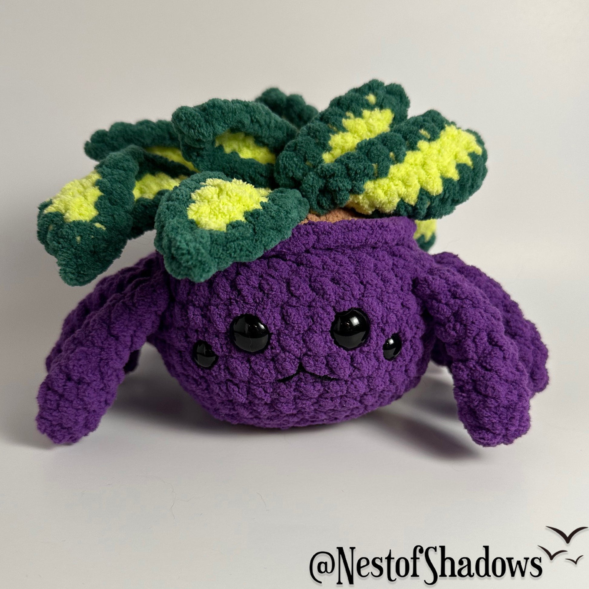 Crochet plushie of a purple spider and plant with a white background and Nest of Shadows watermark.