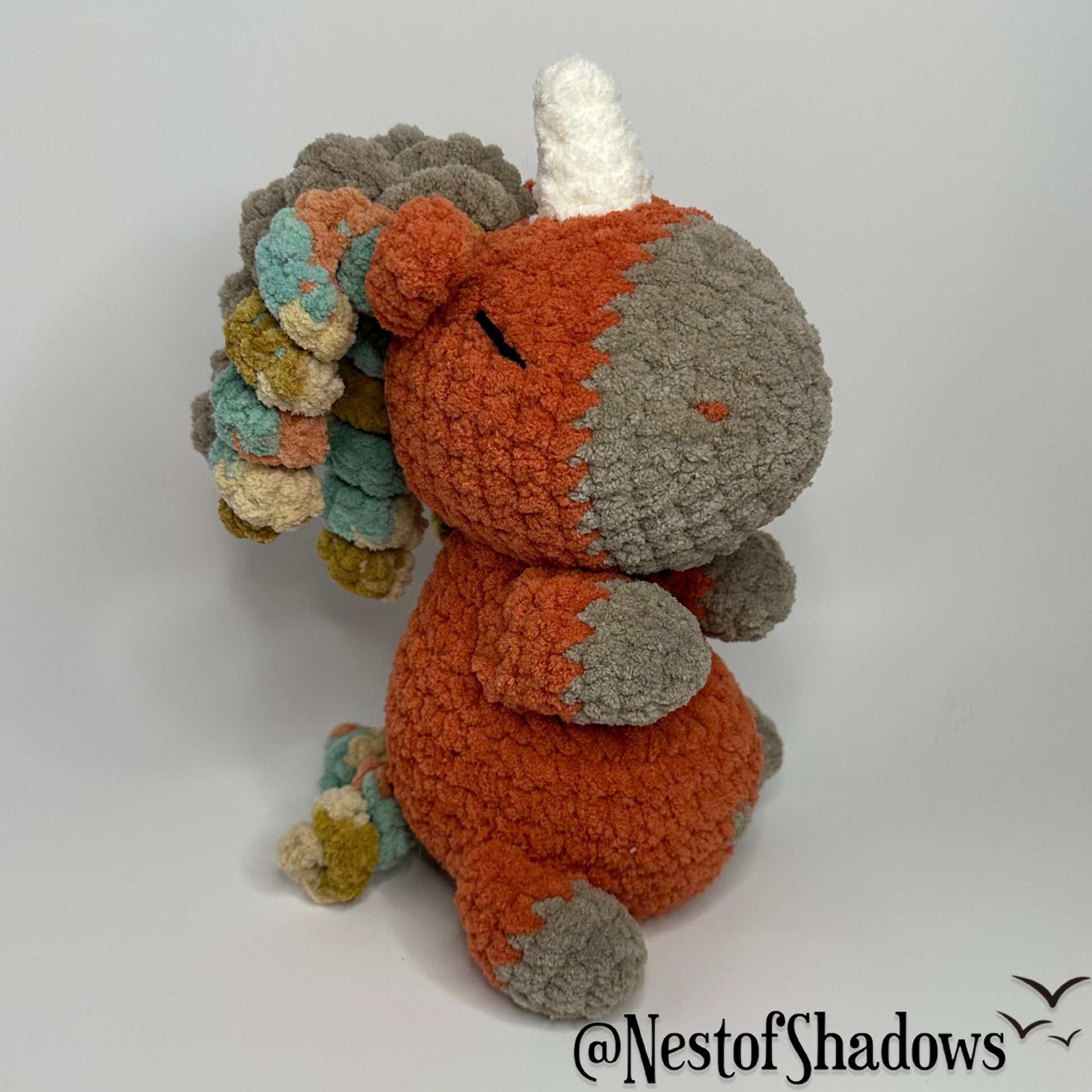 Crochet Unicorn in orange with a white background and a Nest of Shadows watermark.