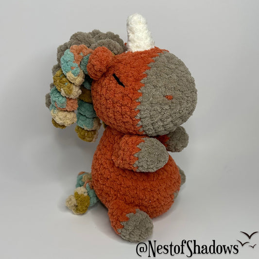 Crochet Unicorn in orange with a white background and a Nest of Shadows watermark.