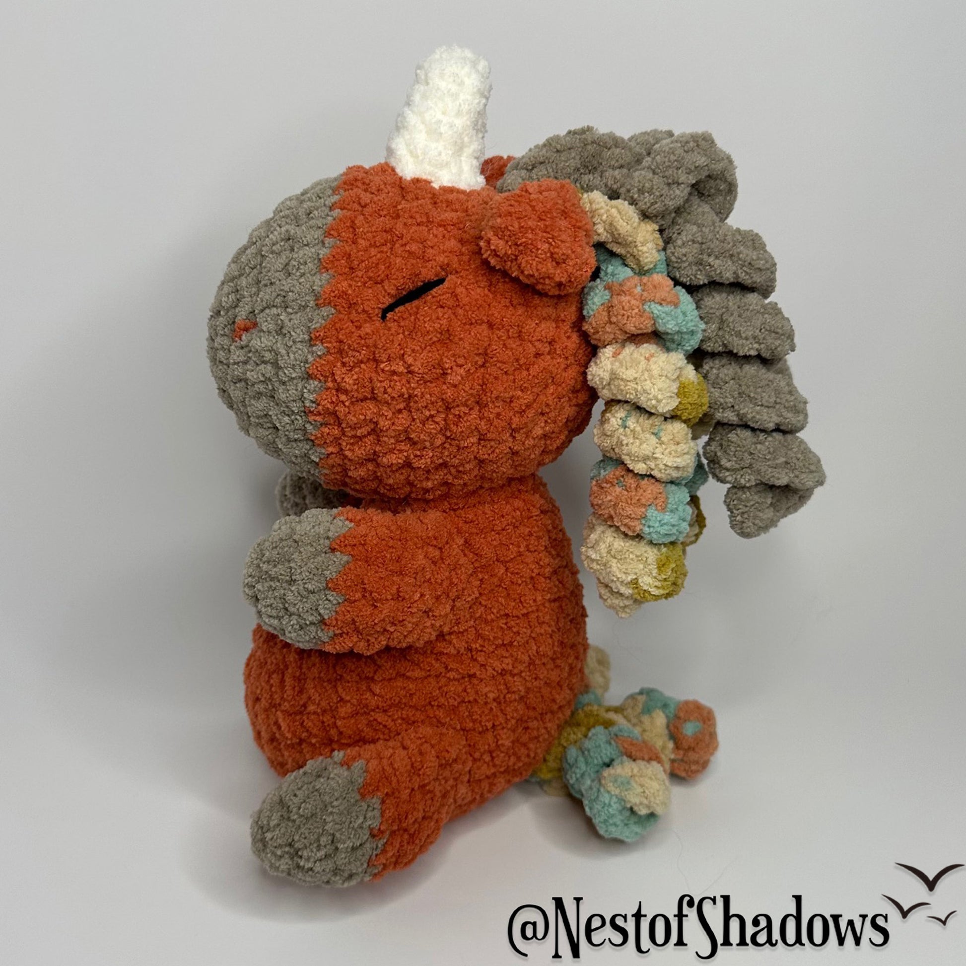 Crochet Unicorn in orange with a white background and a Nest of Shadows watermark.