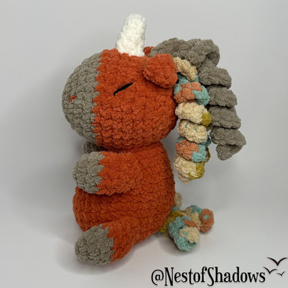 Crochet Unicorn in orange with a white background and a Nest of Shadows watermark.