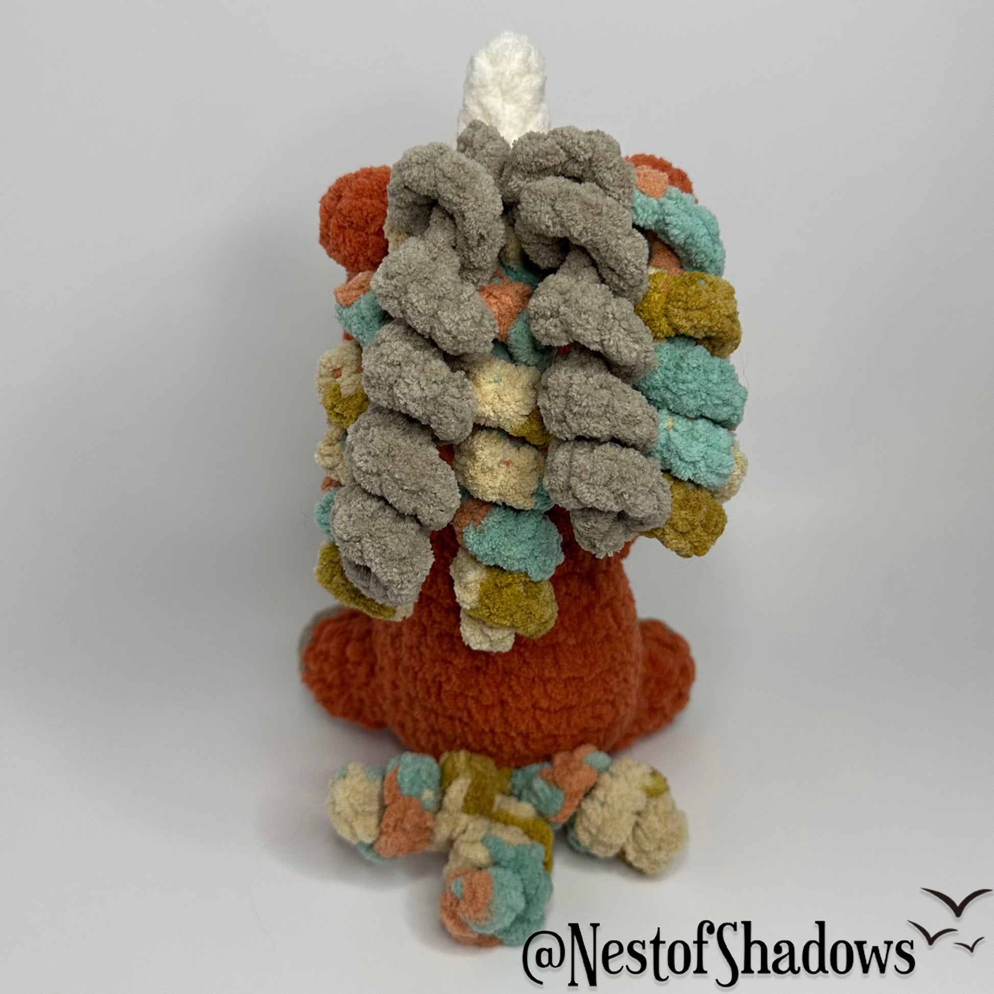 Crochet Unicorn in orange with a white background and a Nest of Shadows watermark.